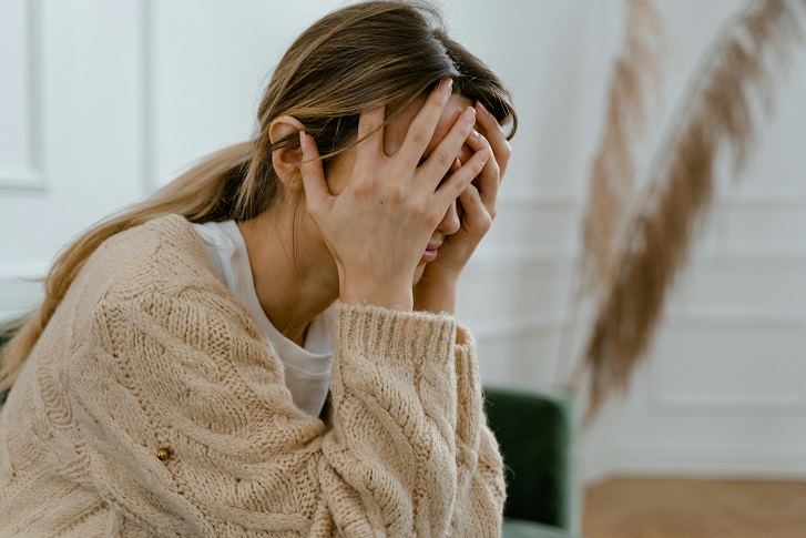 Woman in beige knit sweater covering her face with both hands, appearing stressed or upset indoors.
